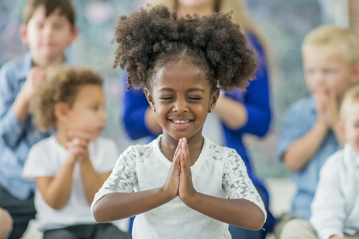 A young Black girl folds her hands in prayer in front of a group of children at a church service. Get A Guide to Mandated Reporter Training for Churches at MandatedReporterTraining.com