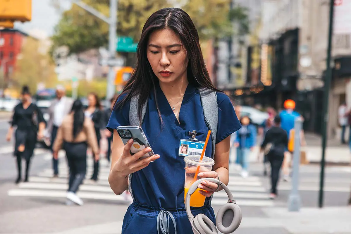 A nurse stands on the street looking at her phone. She is holding a set of headphones and juice.