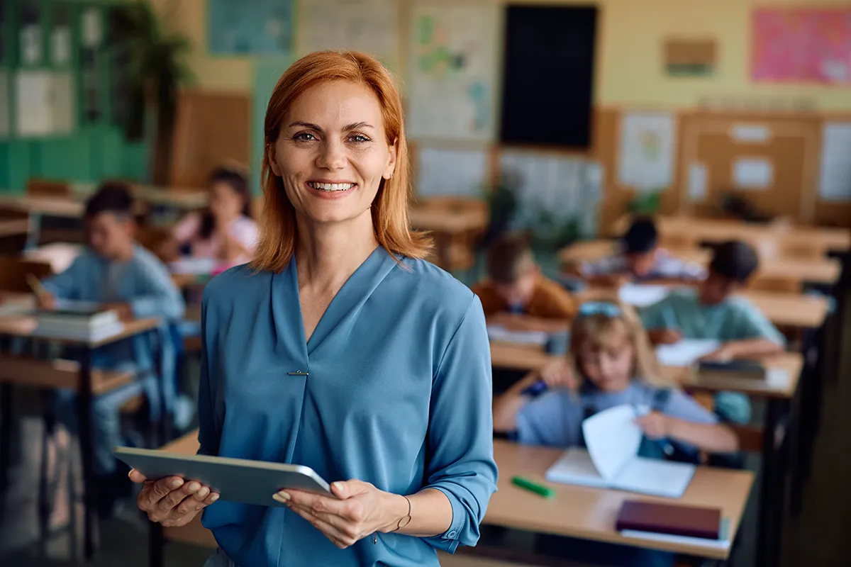 A California teacher wearing a blue blouse, standing in front of a classroom of students. Learn CA AB 1913/ SB 848 training requirements for teachers.