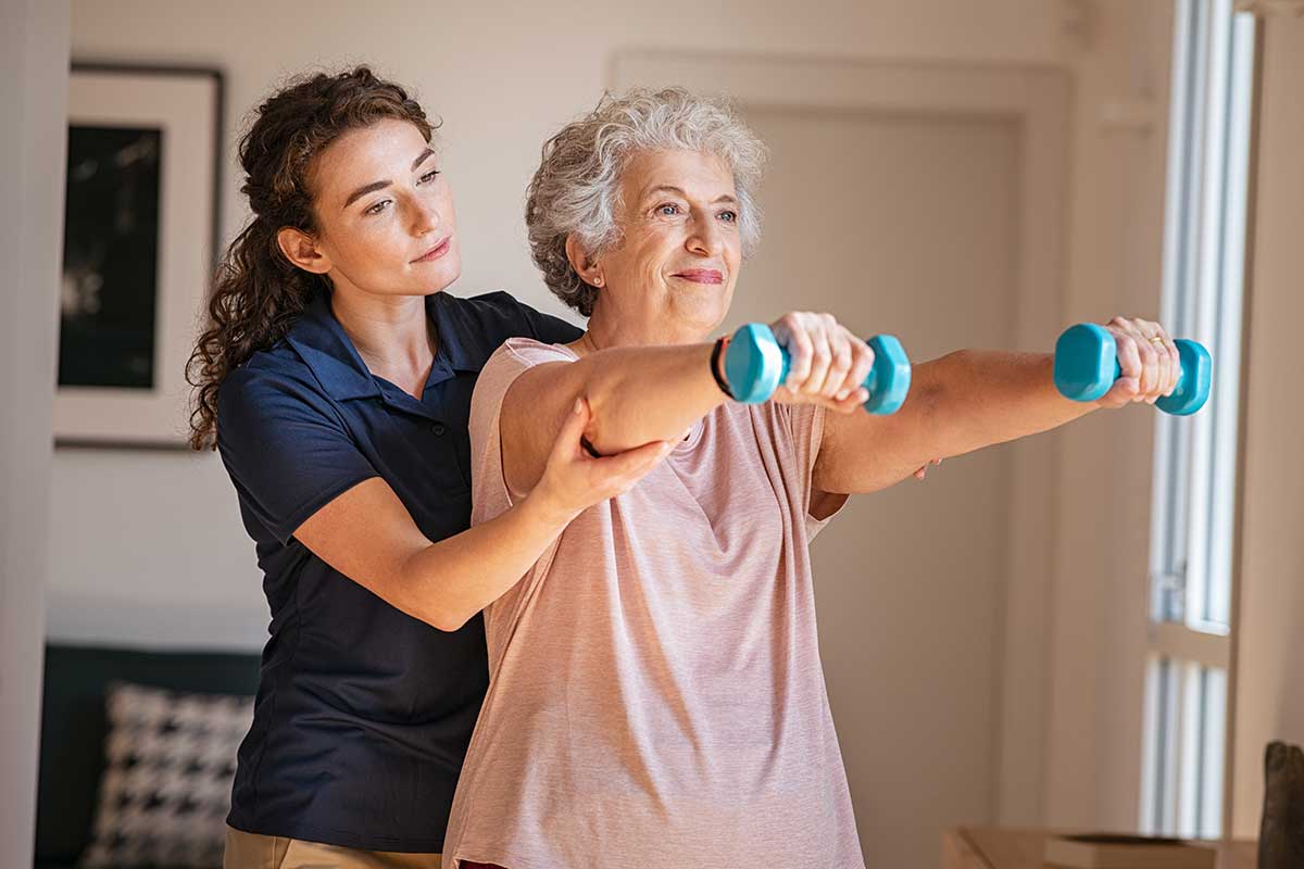 A young female physiotherapist assisting an elderly female client with an arm workout. Find out if elder care businesses need Mandated Reporter Training at our blog.