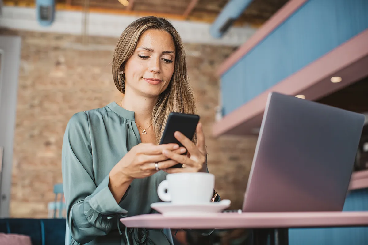 A woman in a cafe holds her phone up to her laptop computer, as if scanning a QR code.