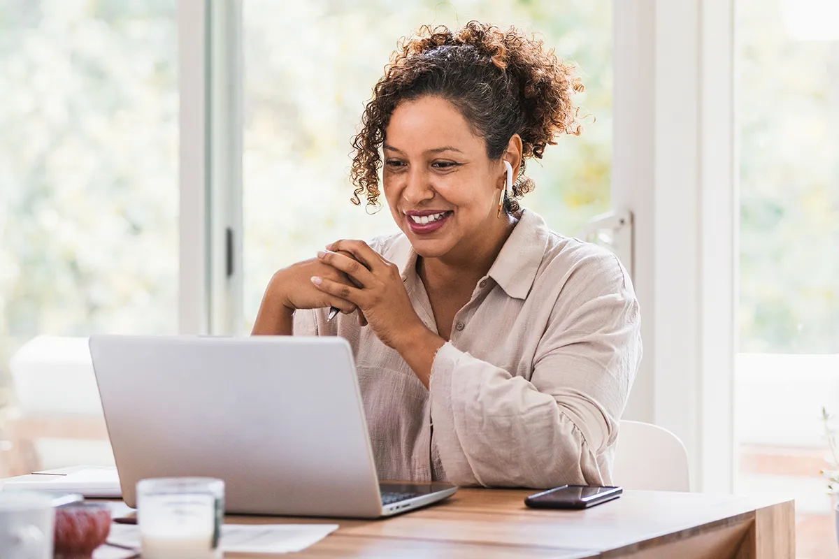 A woman sits at her kitchen table, watching courses on her laptop while wearing earbuds.