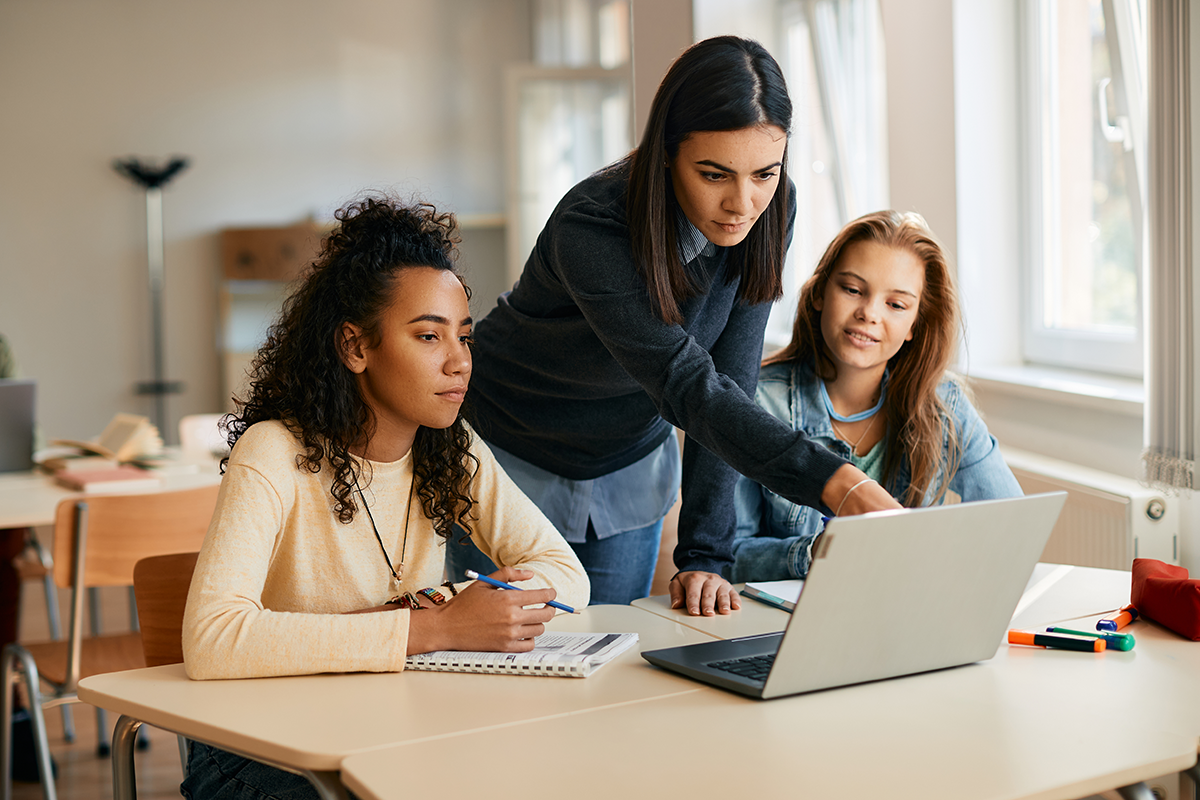 A teacher speaks with two high school students who are using a laptop.