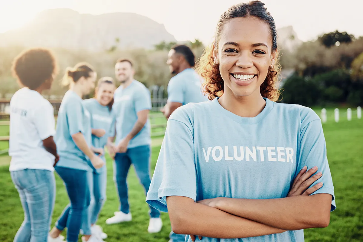 A smiling young Black woman wearing a blue Volunteer shirt stands outside with a volunteer group.