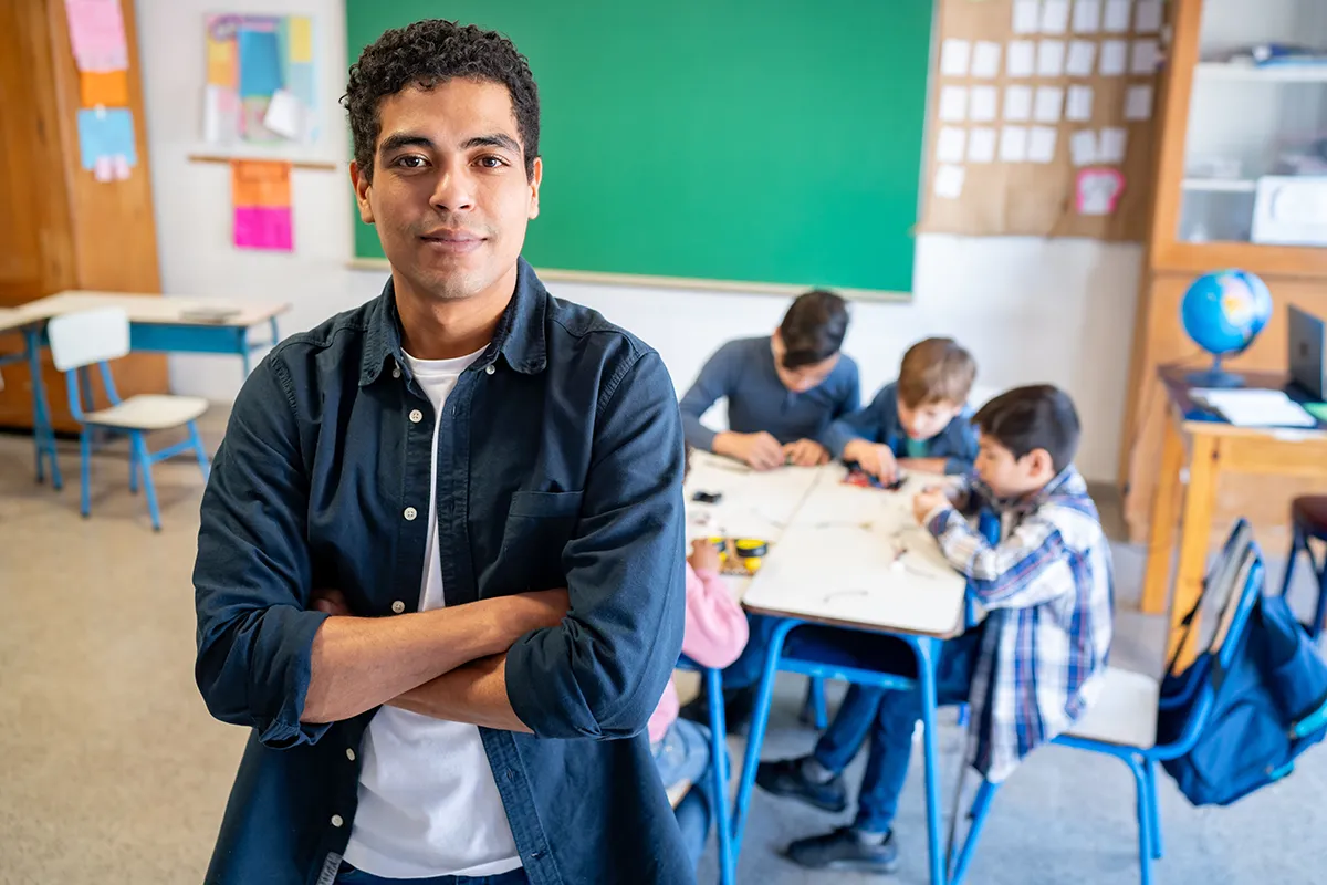 A male teacher stands in front of a classroom with a chalkboard anda table of grade school kids behind him.