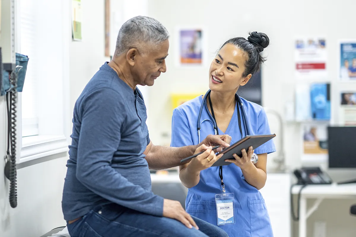 A CA medical professional in a hospital speaks with a patient, showing him something on a clipboard.